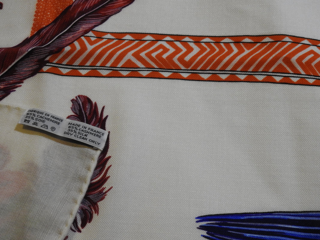 Close up Picture of caretag in used Hermes 140cm silk and cashmere shawl for sale Brazil. White background with bright blue and orange feathers