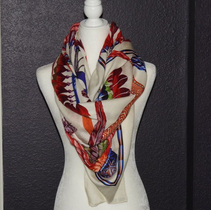 Picture of used Hermes 140cm silk and cashmere scarf for sale. Brazil by Laurence Bourthoumieux has a white background, red, orange, blue, green and purple feathers. This is tied loosely around the neck