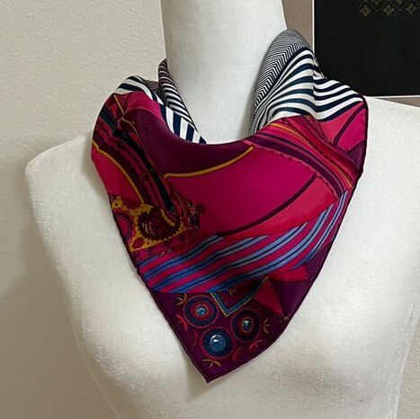 Picture of Coupons Indiens, a used Hermes 45cm silk scarf for sale, knotted in a loose cowboy knot. This scarf is purple and bright pink with pops of turquoise and mustard yellow